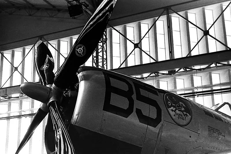 Hélice e fuselagem de um avião antigo com o número "B5", diante das janelas do hangar