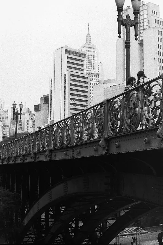 Viaduto de ferro ornamentado com pessoas na calçada e arranha-céus ao fundo
