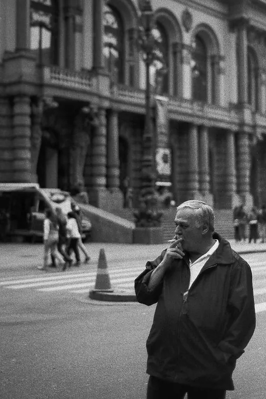 Homem fumando, de jaqueta escura, em uma rua diante de um grande edifício histórico