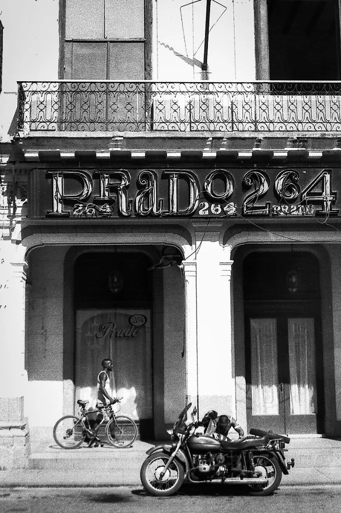 Fachada com letreiro "Prado 264" e sacada de ferro; um ciclista e uma moto com sidecar estacionada na calçada