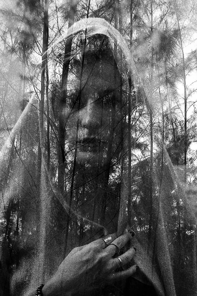 Dupla exposição: retrato de mulher coberta por um tecido translúcido sobreposto a uma floresta de pinheiros